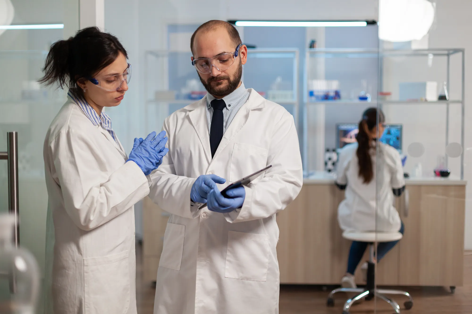 Dos personas con delantal blanco, lentes de protección transparente y guantes azules mirando un registro en un laboratorio.