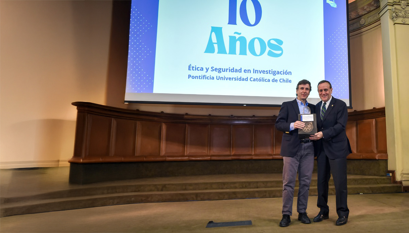 Dos personas, Rector de la Pontificia Universidad Católica de Chile, Sr Ignacio Sánchez entregando un reconocimiento a Juan Larraín académico de la misma universidad en el salón Aula Magna de la universidad. En el escenario la bandera del Vaticano y la bandera de Chile.